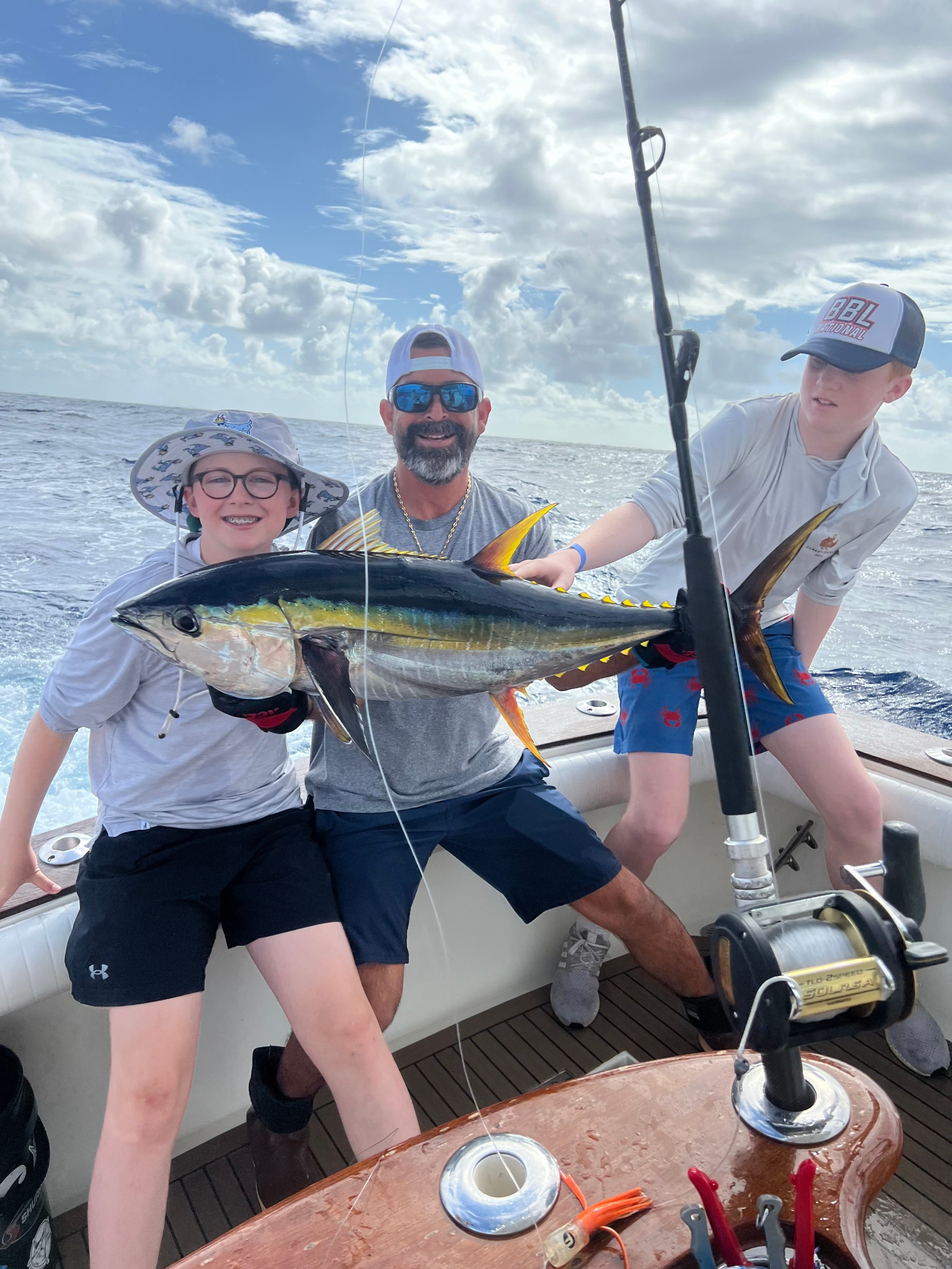 Three people on a boat proudly holding a large fish with fishing rods in view, under a cloudy sky.