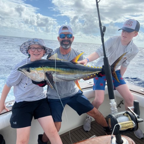 Three people on a boat proudly holding a large fish with fishing rods in view, under a cloudy sky.