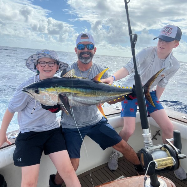 Three people on a boat proudly holding a large fish with fishing rods in view, under a cloudy sky.