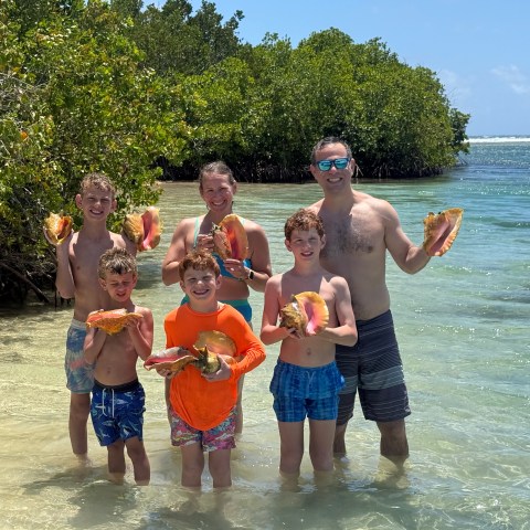 Family holding conch shells on a beach by clear water and trees.