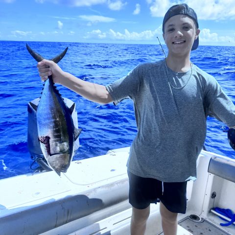 Person smiling, holding a large fish on a boat with blue ocean and sky.