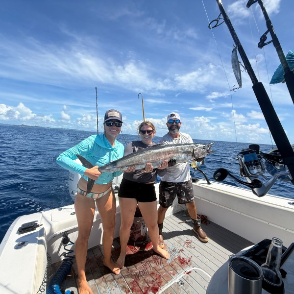 Three people on a boat holding a large fish under a blue sky.