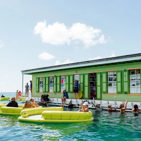 People gather around a floating green bar with yellow floats in the ocean under a blue sky.