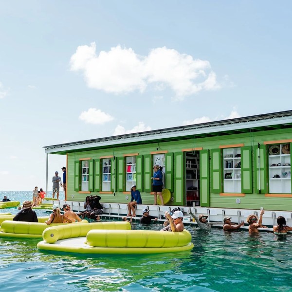 People gather around a floating green bar with yellow floats in the ocean under a blue sky.